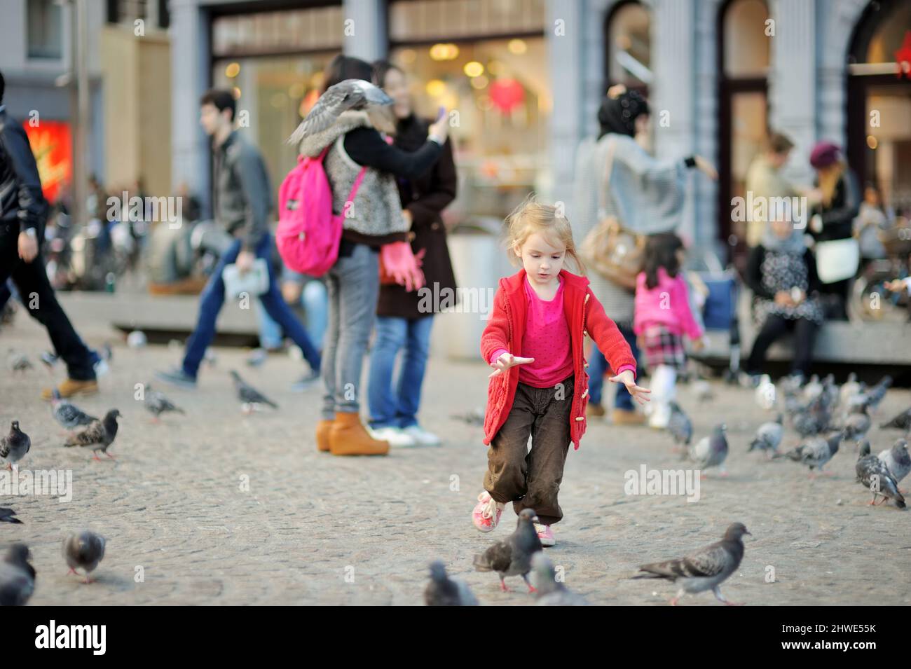 Carina bambina che allena e insegue gli uccelli in piazza Dam ad Amsterdam il giorno d'estate. Bambini che allattano piccioni e passeri all'aperto. Foto Stock