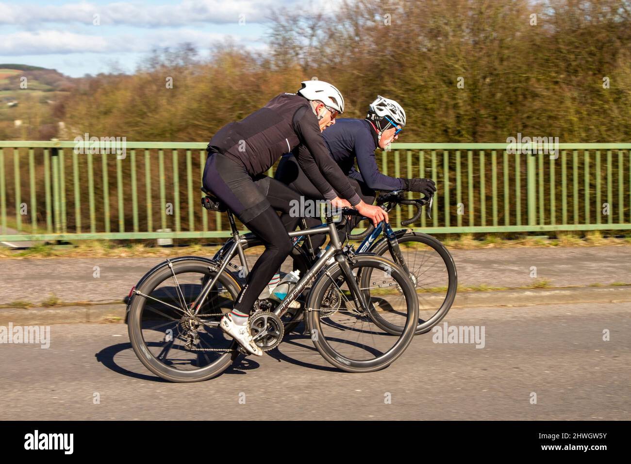Due ciclisti maschi che cavalcano RaceLight & Trek sport Road bike, affiancati, sulla strada di campagna che attraversa il ponte autostradale nella campagna del Lancashire, Regno Unito Foto Stock