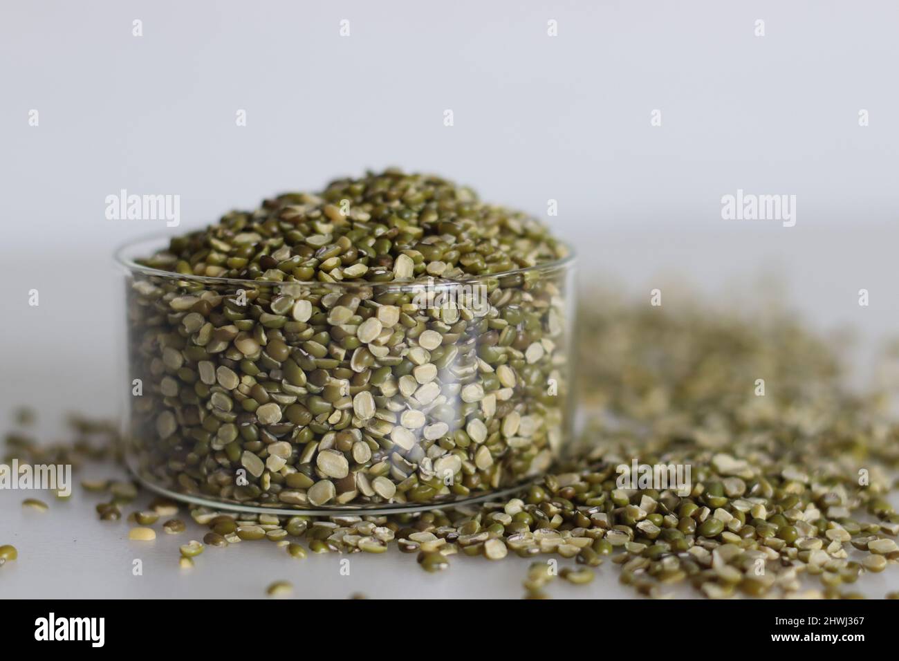 Dividere le lenticchie di fagiolo di moong in una piccola ciotola di vetro. Sparato su sfondo bianco con mucchio di fagioli di moong divisi sono sparsi intorno. Foto Stock