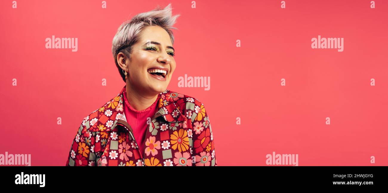 Donna allegra hipster che guarda via con un sorriso sul suo volto in uno studio. Giovane donna felice in piedi da sola su sfondo rosso. Giovane alla moda Foto Stock