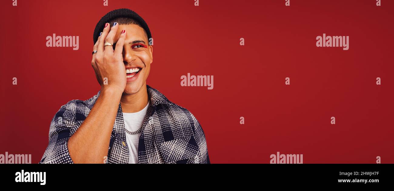 Giovane uomo non conforme che indossa smalto per unghie e trucco in uno studio. Felice giovane uomo sorridente allegro mentre si alza da solo su sfondo rosso. Foto Stock