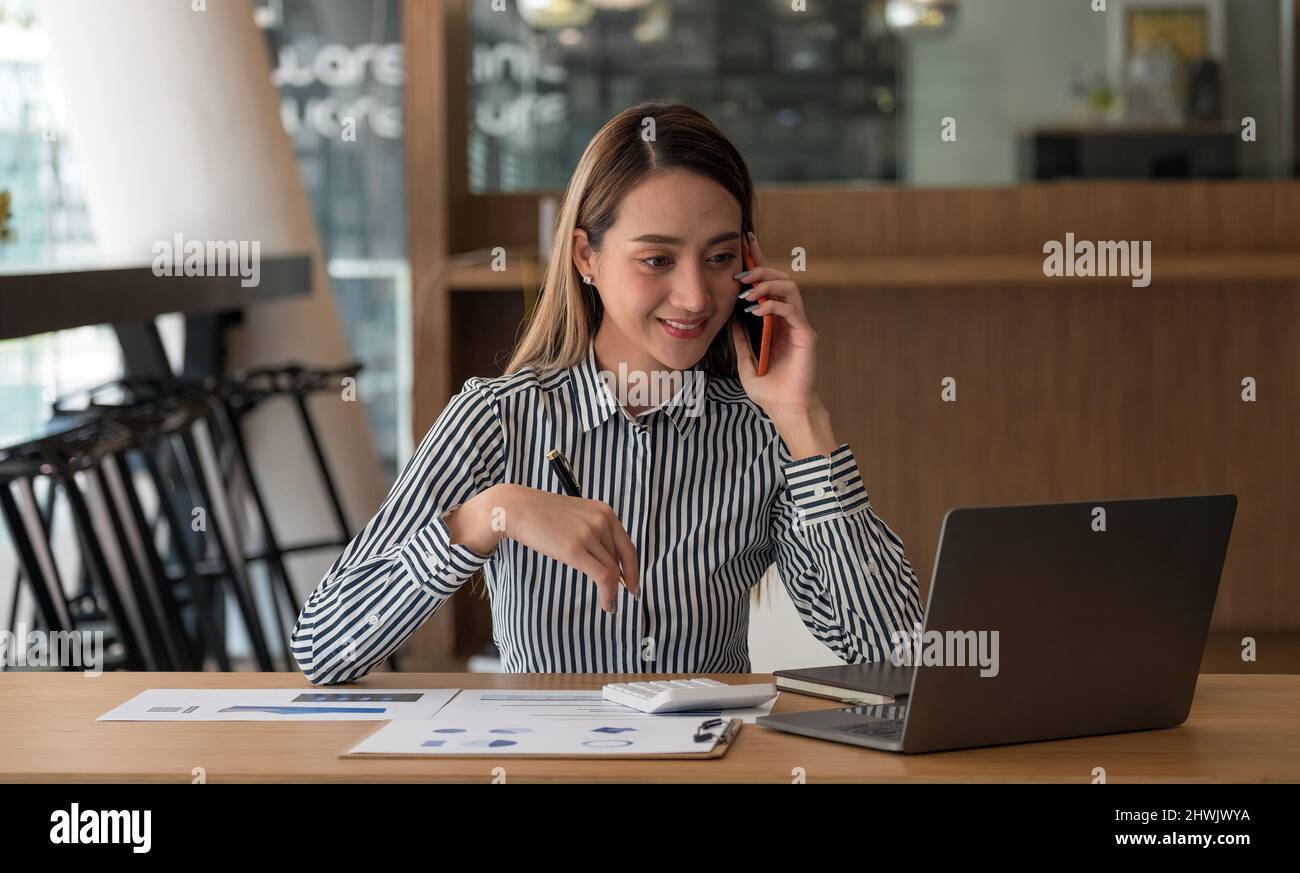 Immagine dell'affascinante donna asiatica che parla con lo smartphone e lavora su un computer portatile, mentre si siede al tavolo al coperto. Foto Stock