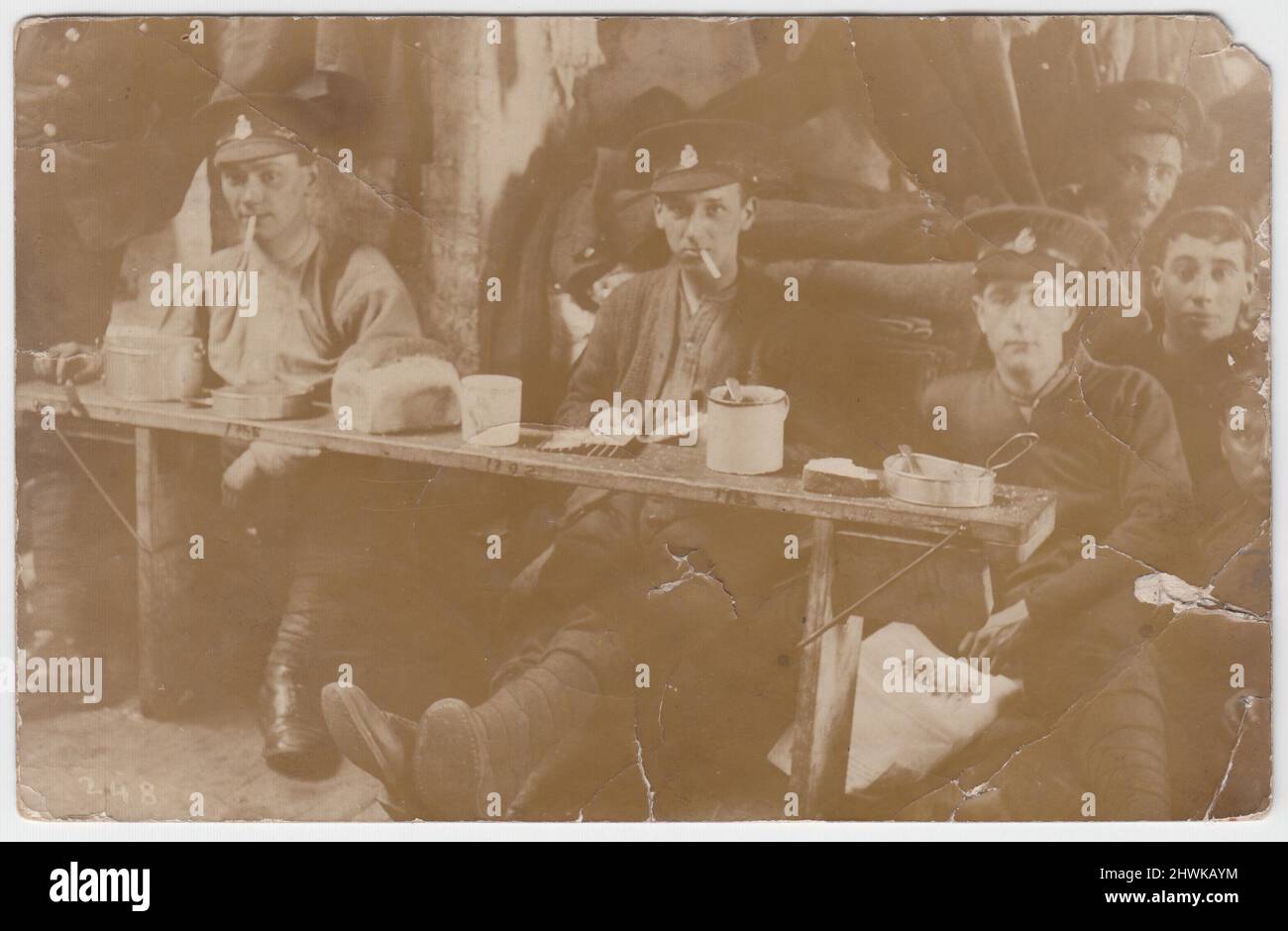 Fotografia informale dei soldati britannici della prima guerra mondiale che mangiano razioni. I giovani sono seduti a una panca bassa o a un tavolo che ha pane, tini di mess & tazze di stagno sopra. Due sigarette fumano e una ha un giornale in grembo, un soldato sembra indossare un cardigan, oltre alla sua uniforme militare standard. L'alloggio sembra temporaneo, forse un fienile o un altro edificio sul fronte occidentale Foto Stock