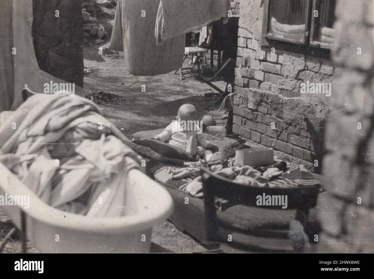 antica foto monocromatica su un bambino carino bambino che è seduto nel suo parco giochi e giocare in un povero condizionata (non comfort) casa giardino. Le pareti in mattoni sono visibili della casa, il bagno è all'esterno e i vestiti e le sedie sono anche lonside. Sembra un deposito di rottami o che era il normale dopo il periodo della prima guerra mondiale: 1920s . Fonte: Fotografia originale. ULTERIORI-DIRITTI-SPAZIO-INFORMAZIONI-NON-DISPONIBILI Foto Stock