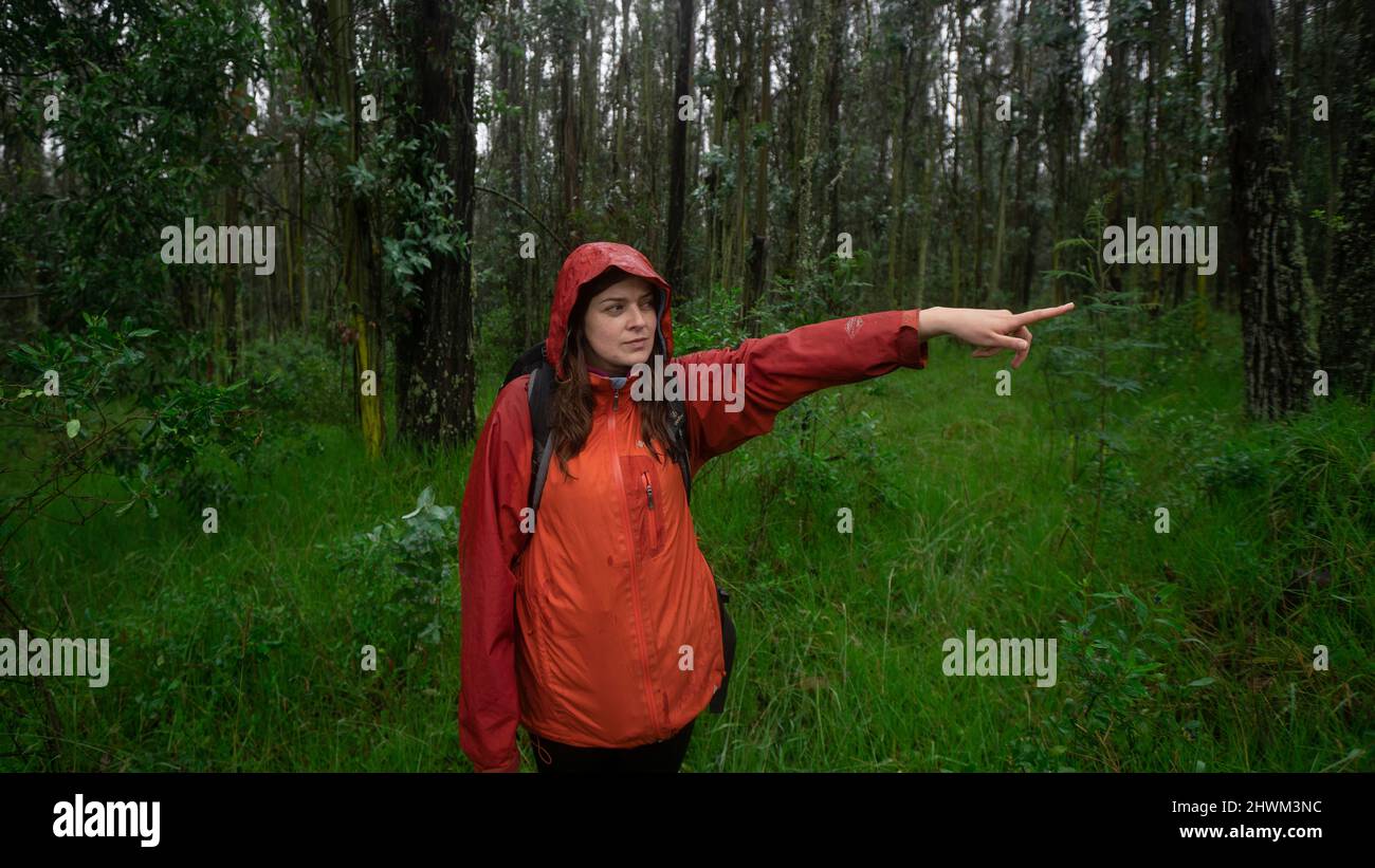 Vista frontale di una donna ispanica con uno zaino nero e una giacca impermeabile rossa che punta la direzione con il suo braccio allungato al centro di una f Foto Stock