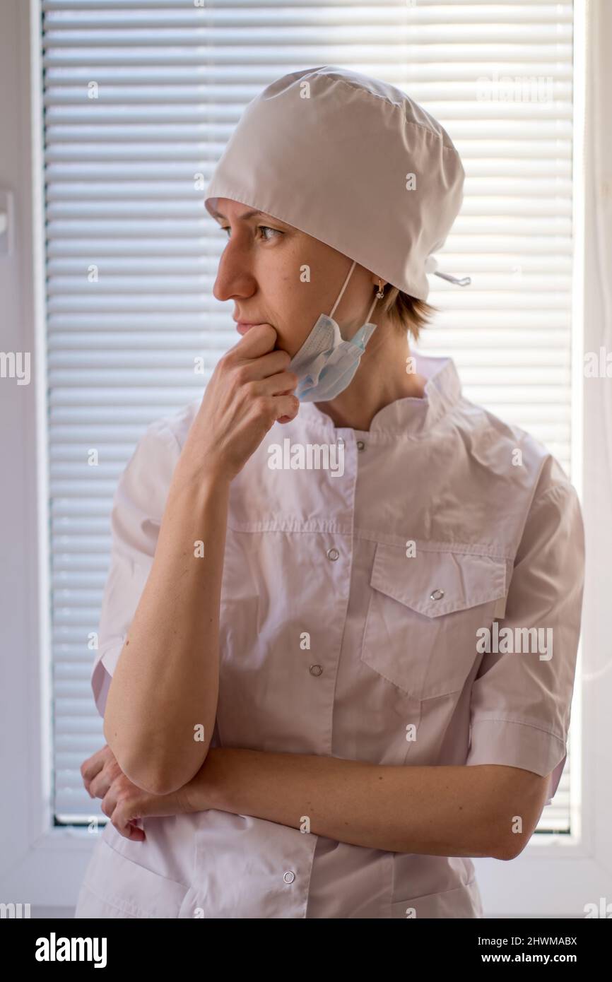 Una dottore femminile con una maschera sul viso si alza in piedi in modo pensivo alla finestra. Foto Stock