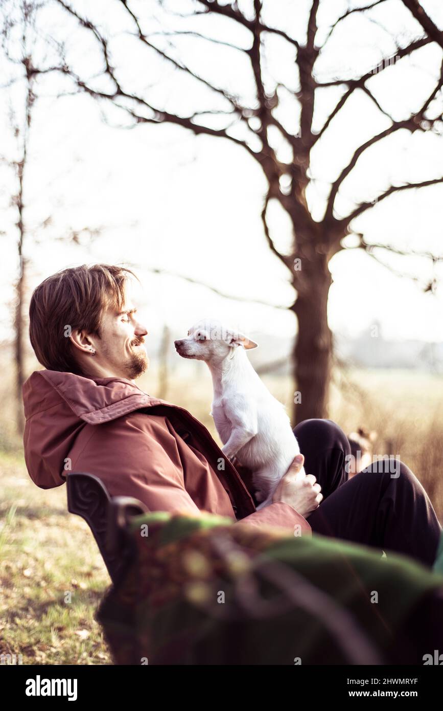 Il giovane felice con giacca si siede all'aperto con il cane in grembo sotto il sole Foto Stock