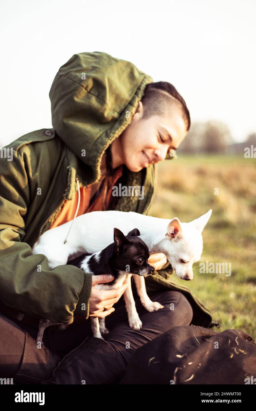 persona androgina si siede in campo soleggiato con due chihuahuas sorridente Foto Stock