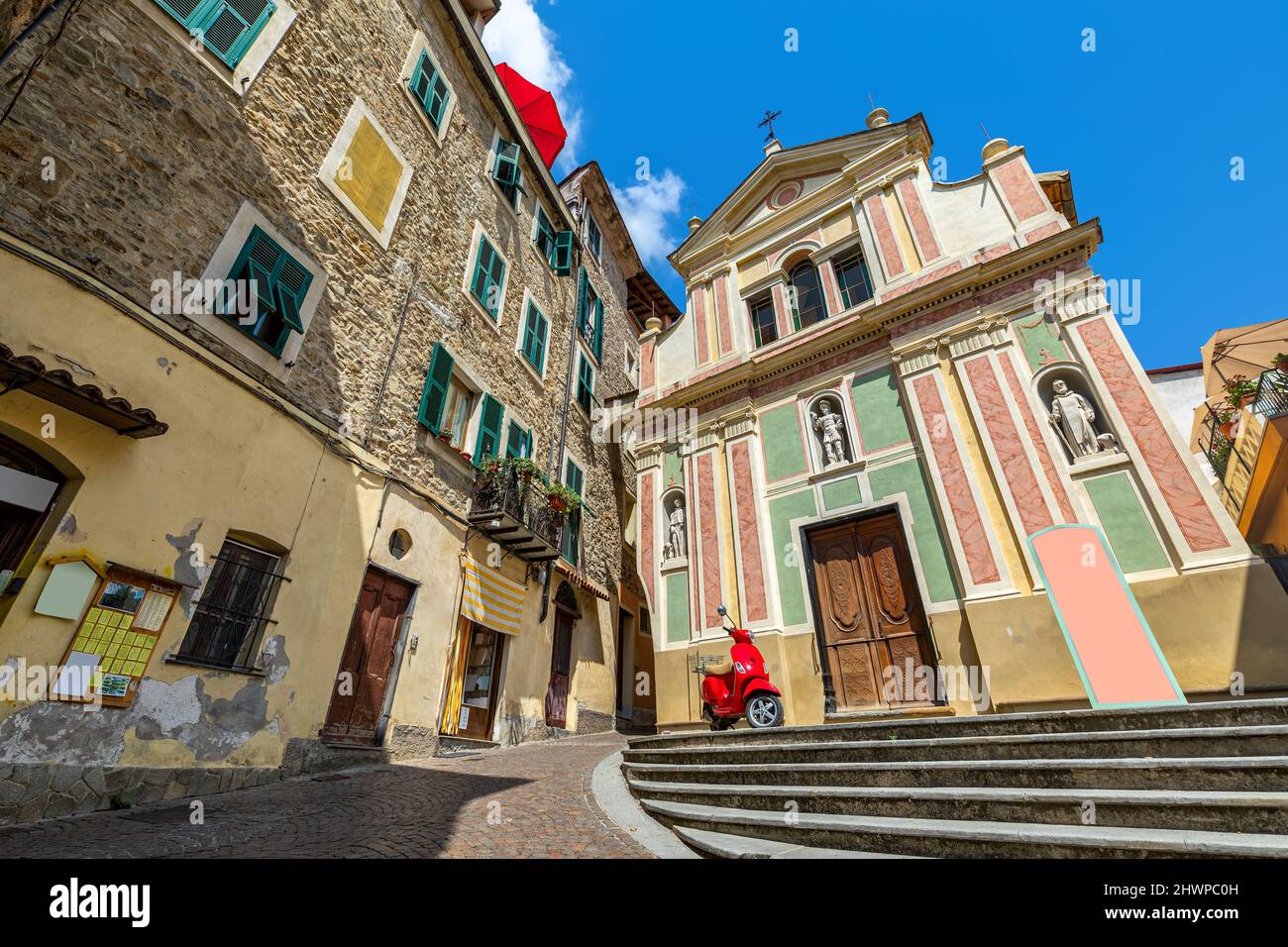 Scooter rosso sulla piccola piazza tra vecchie case in pietra e chiesa cattolica sotto il cielo blu a Dolceacqua, Liguria, Italia. Foto Stock