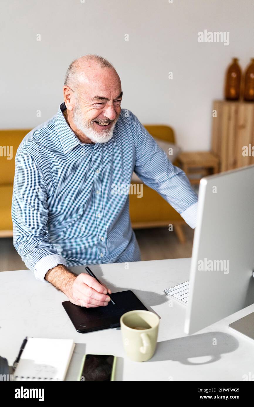 Uomo d'affari anziano allegro con un tablet grafico che guarda il computer in ufficio domestico Foto Stock