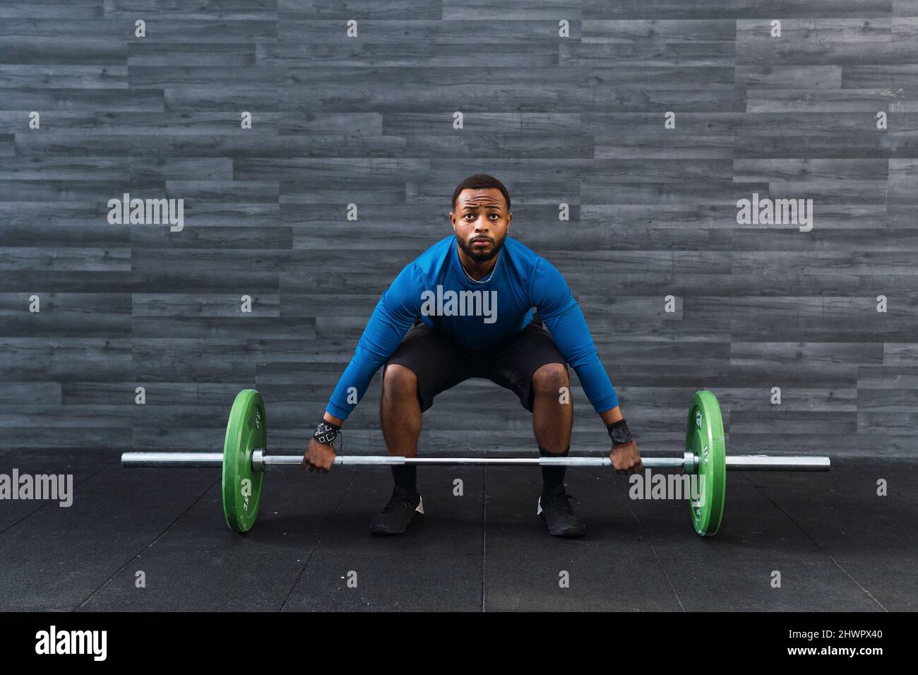 Un giovane atleta concentrato che raccoglie il barbell davanti al muro in palestra Foto Stock