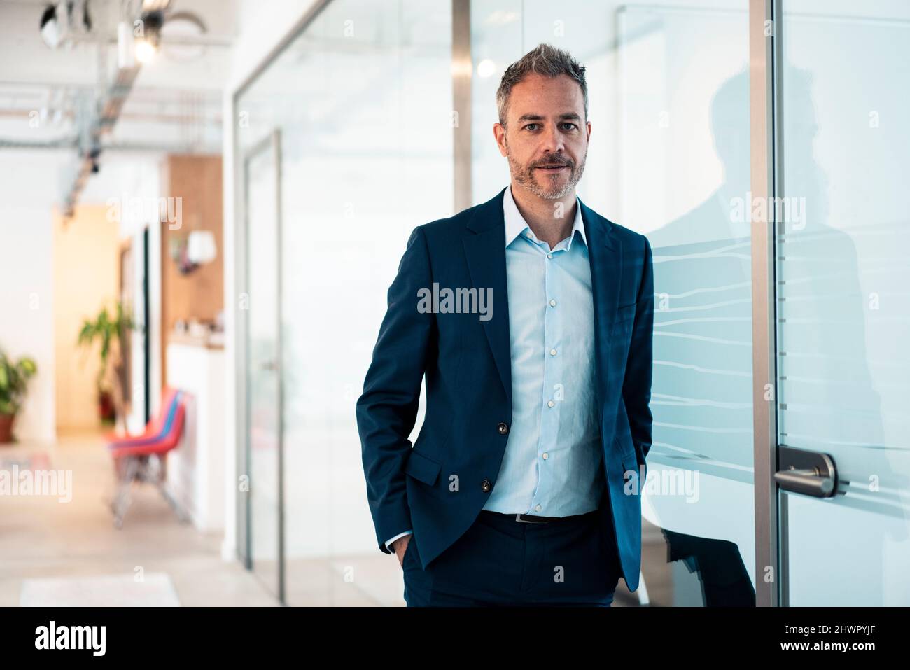 Uomo d'affari sicuro con le mani in tasca appoggiate su una parete di vetro sul posto di lavoro Foto Stock