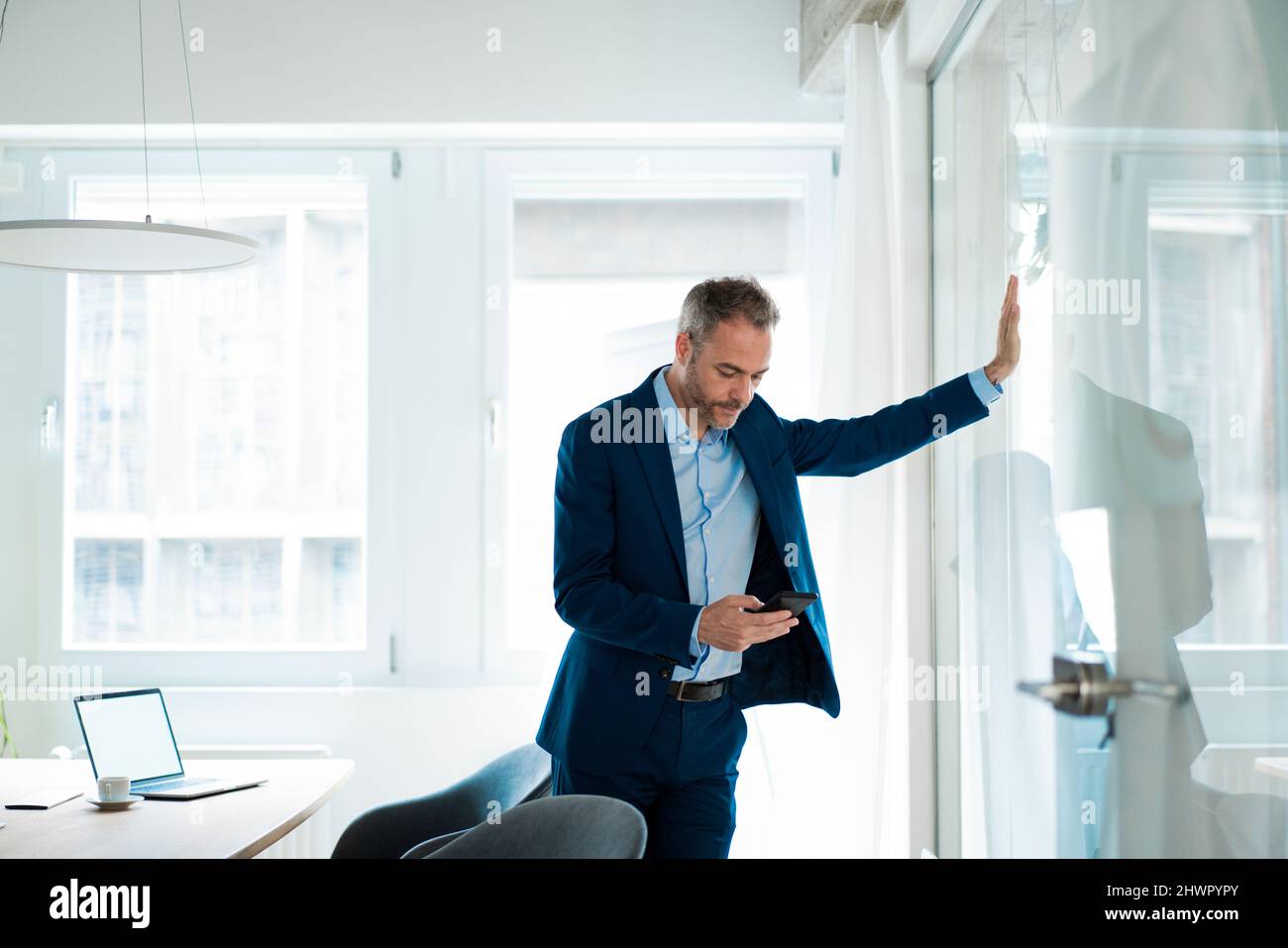 Uomo d'affari nella sala riunioni appoggiata su una parete di vetro messaggi di testo tramite smartphone Foto Stock