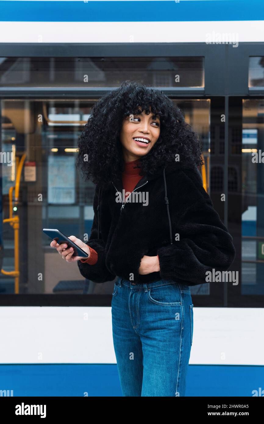 Giovane donna sorridente con smartphone di fronte al tram Foto Stock