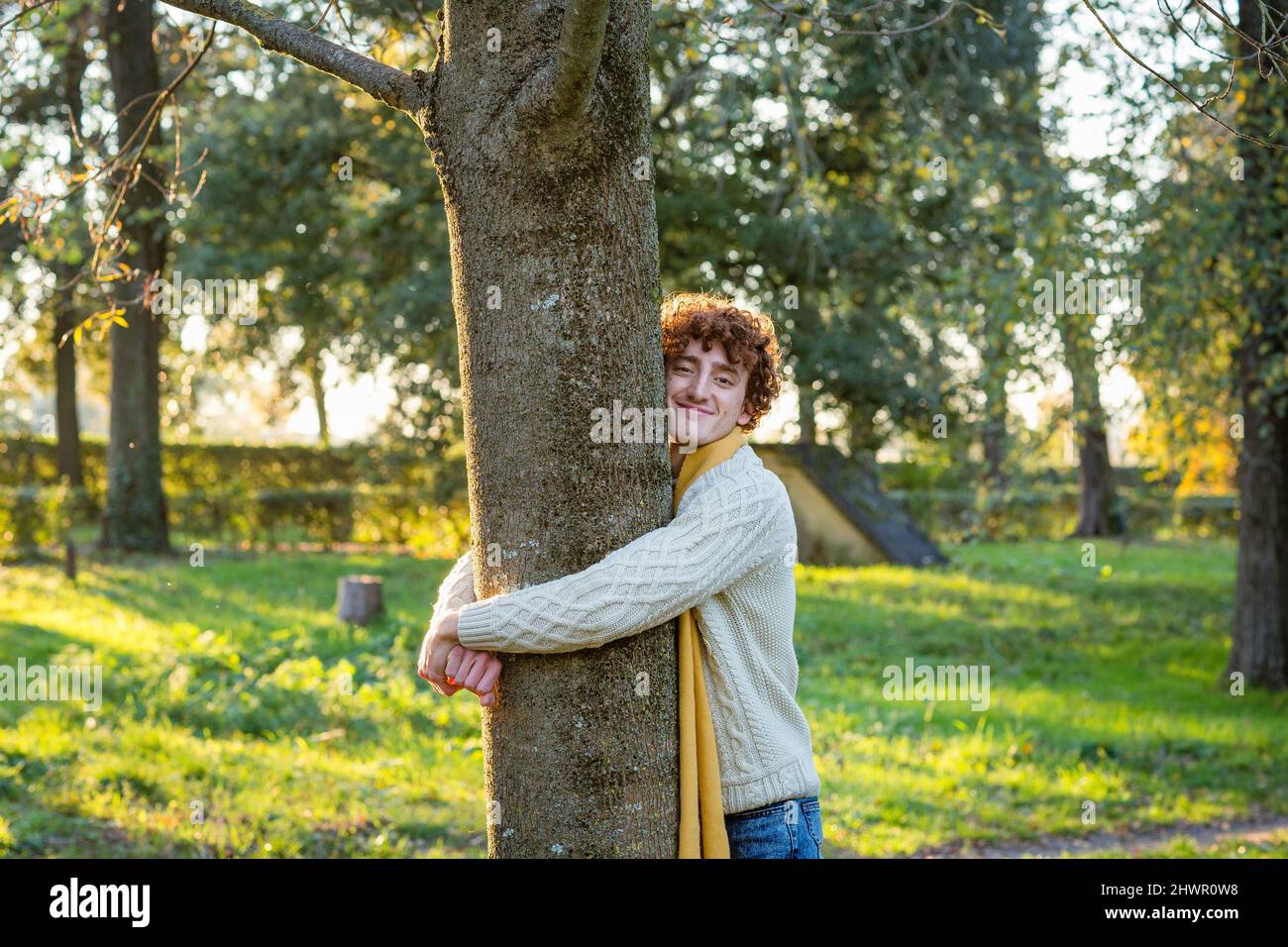 Sorridente giovane uomo che abbraccia l'albero al parco Foto Stock