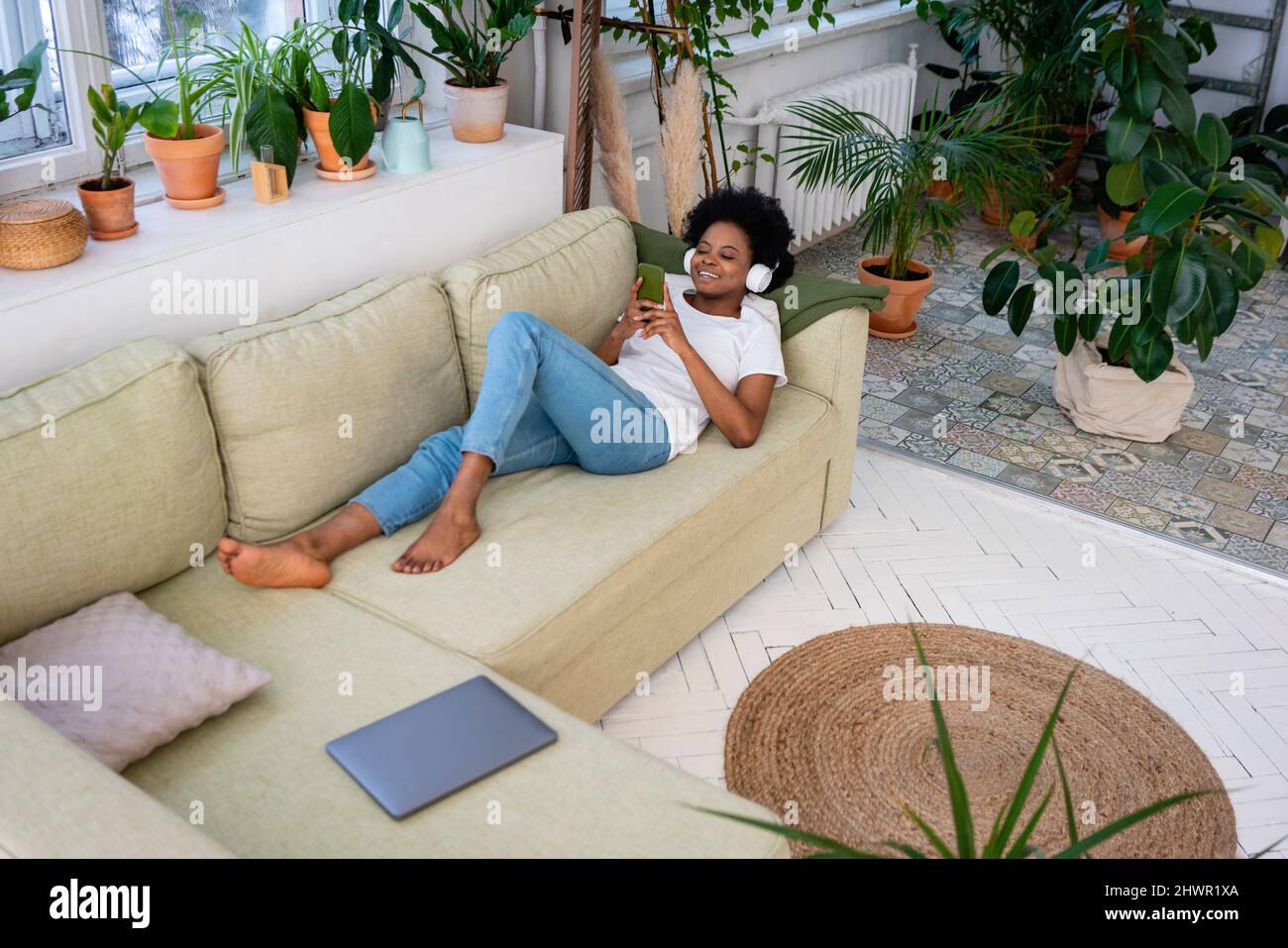 Donna sorridente che utilizza lo smartphone e ascolta la musica tramite le cuffie sul divano di casa Foto Stock