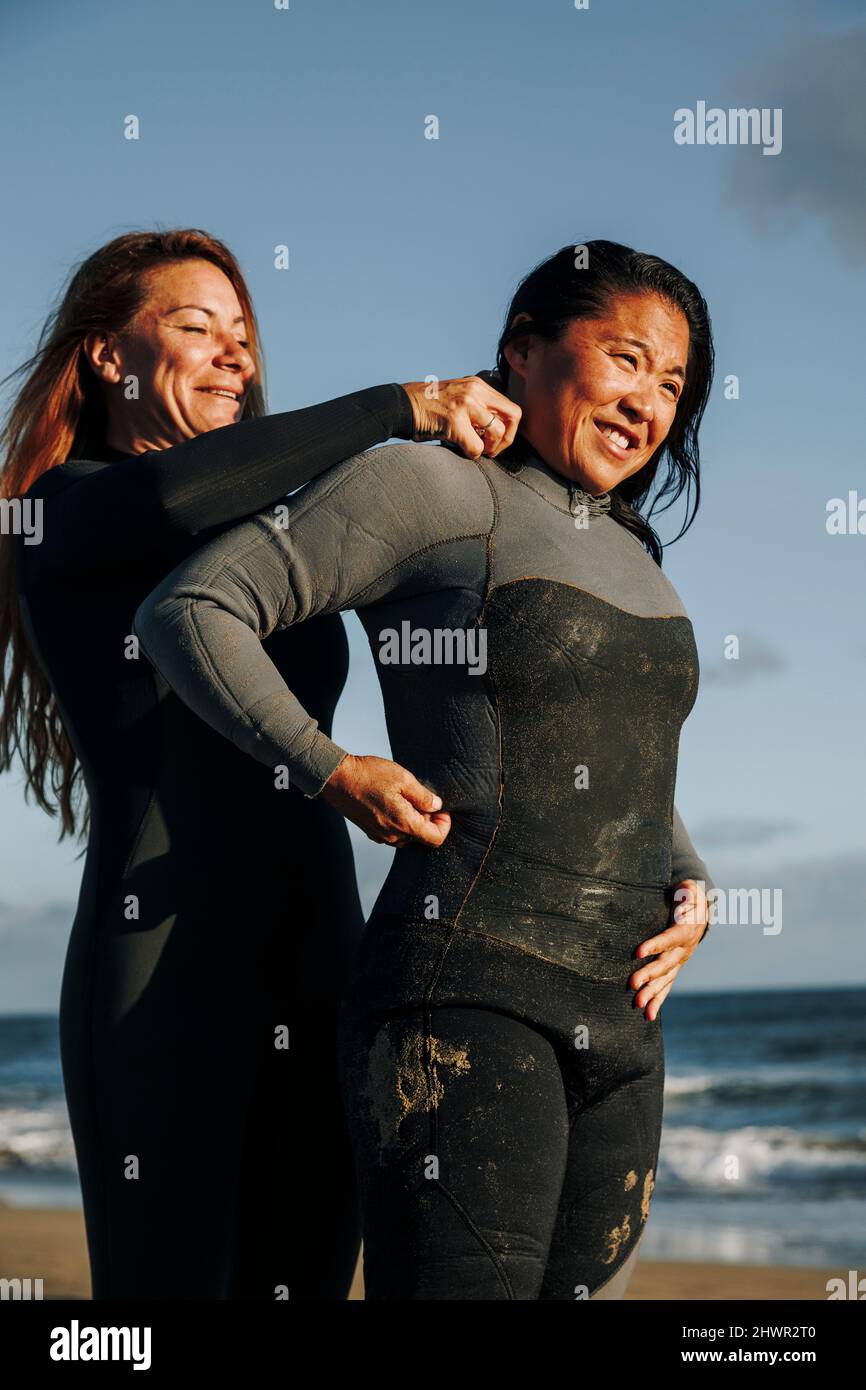 Sorridente surfista che aiuta la donna a mettere la muta in spiaggia Foto Stock