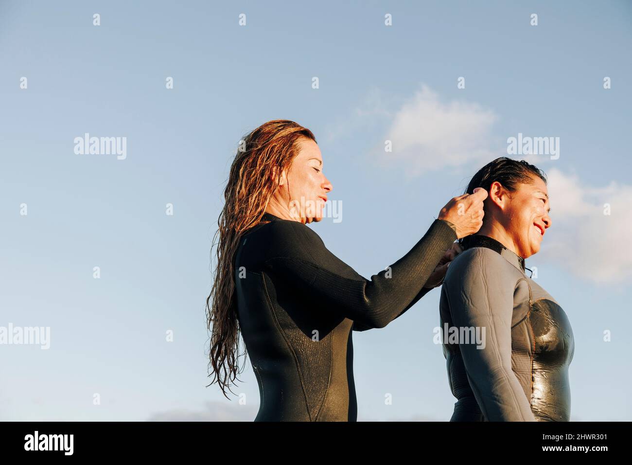 Donna sorridente che aiuta il surfista a mettere la muta in spiaggia Foto Stock