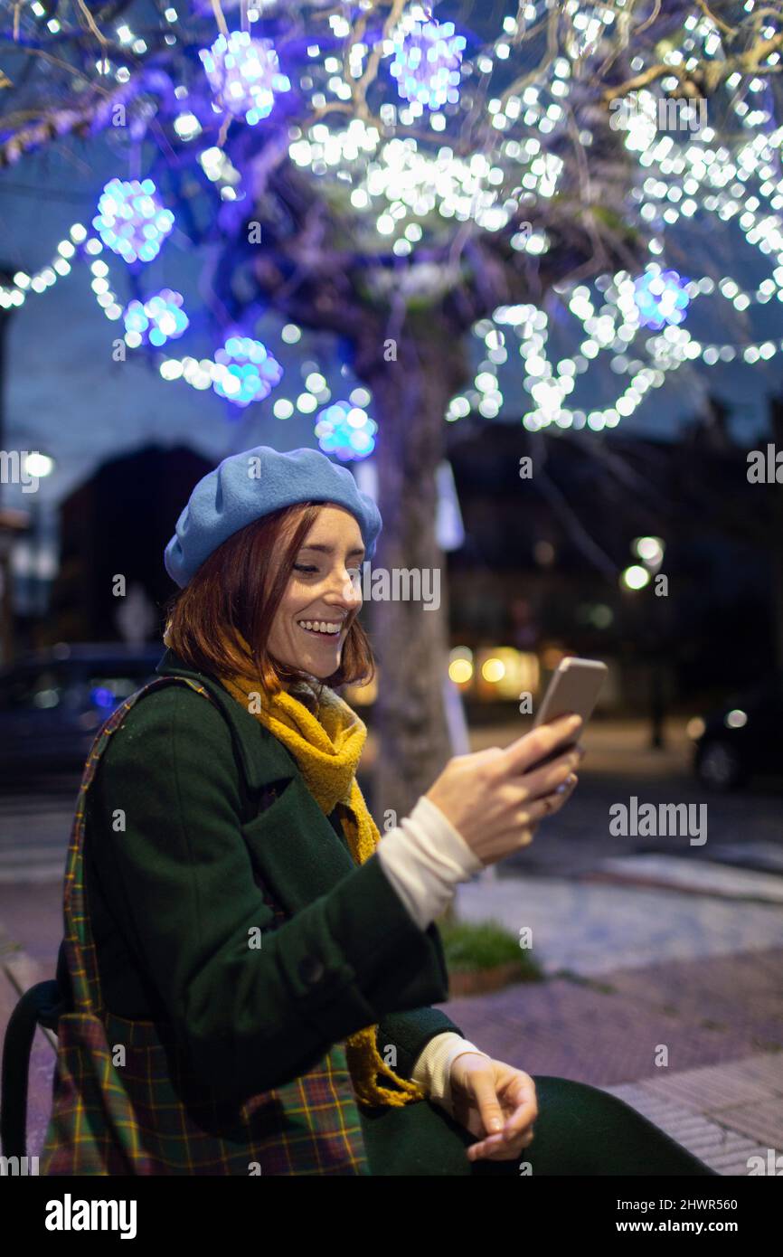Messaggi di testo di una donna felice tramite smartphone seduti di fronte all'albero illuminato di notte Foto Stock