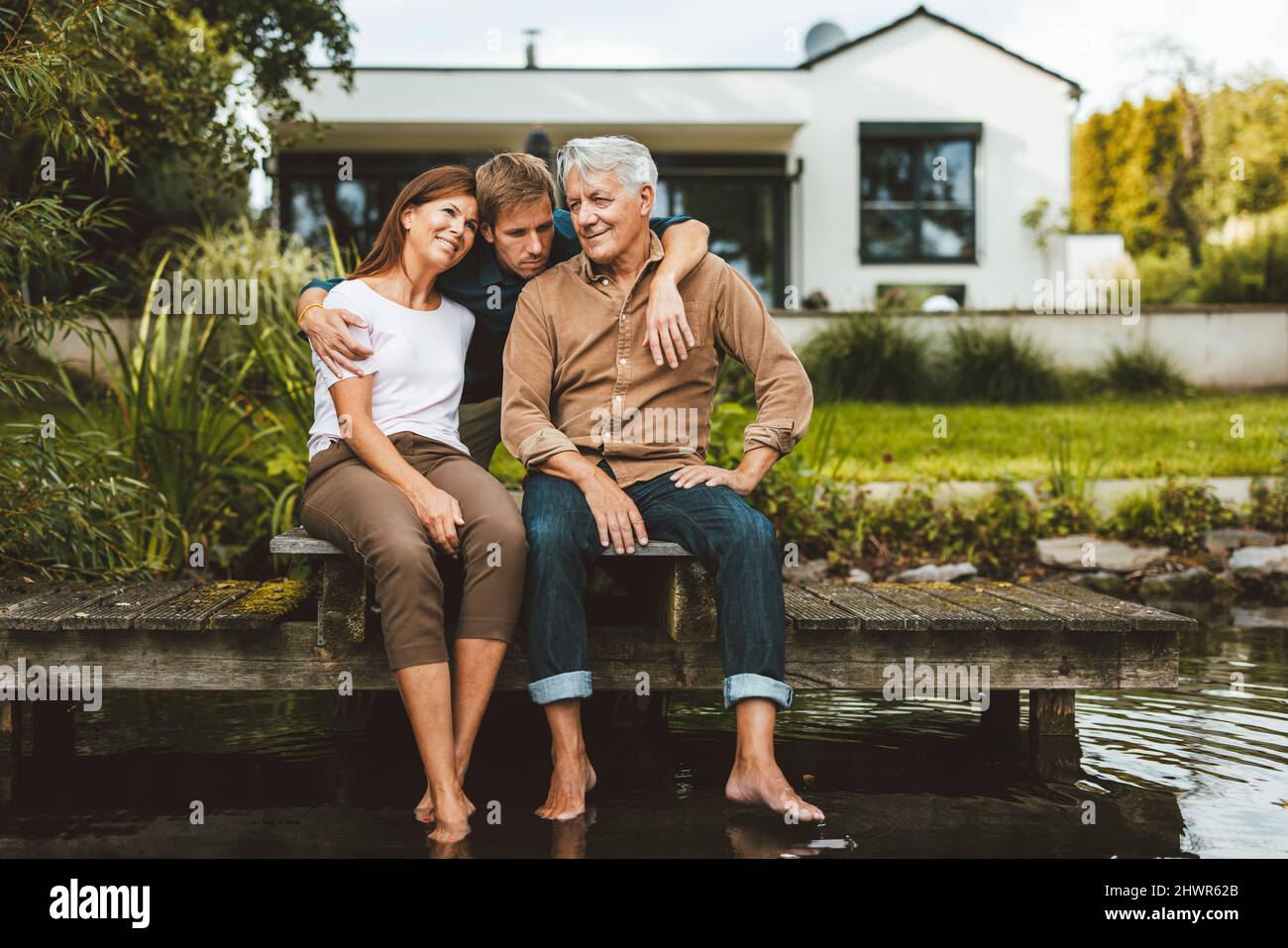 Figlio che abbraccia padre e madre seduto sul molo vicino al lago sul cortile Foto Stock