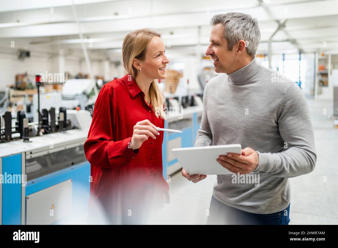 Felice donna d'affari bionda che condivide idee con un collega che tiene un tablet PC in fabbrica Foto Stock