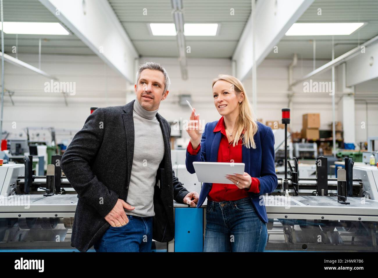 Una donna d'affari sorridente che tiene un tablet PC condividendo idee con un collega che si trova accanto a un macchinario Foto Stock