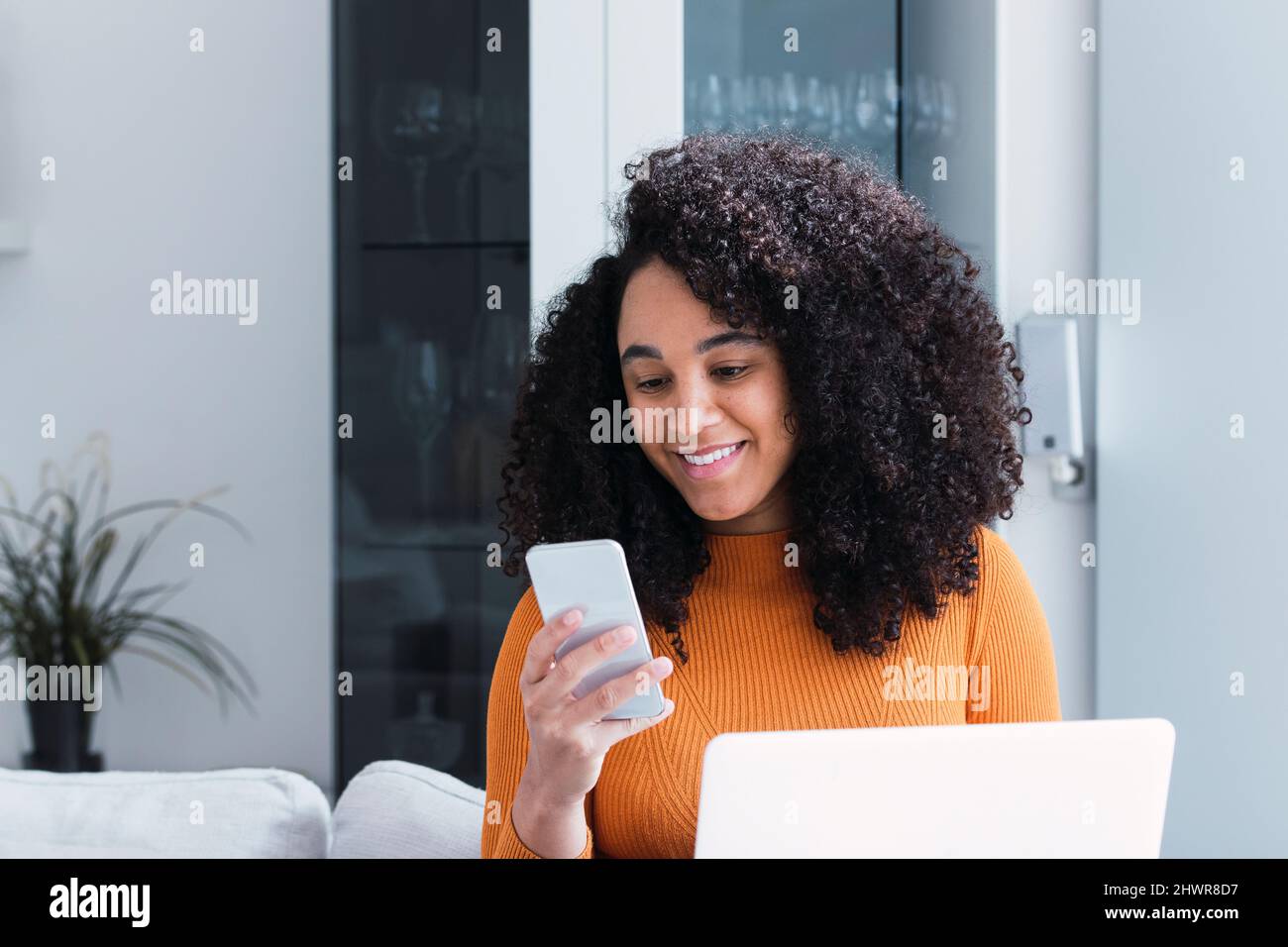 Donna con notebook che utilizza uno smartphone in salotto Foto Stock