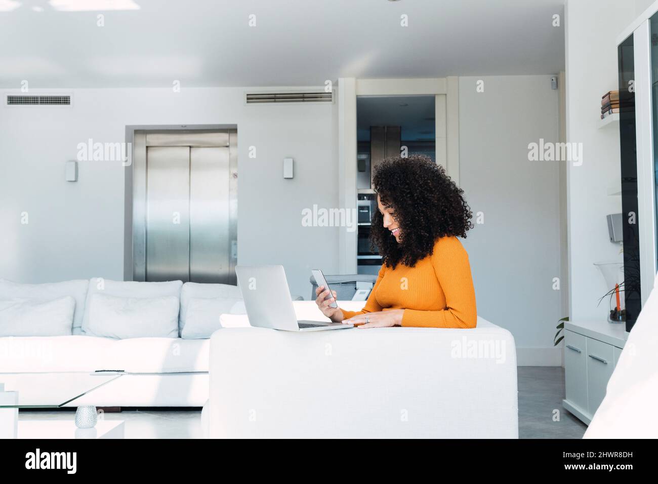 Donna che usa il telefono cellulare da un computer portatile in salotto Foto Stock