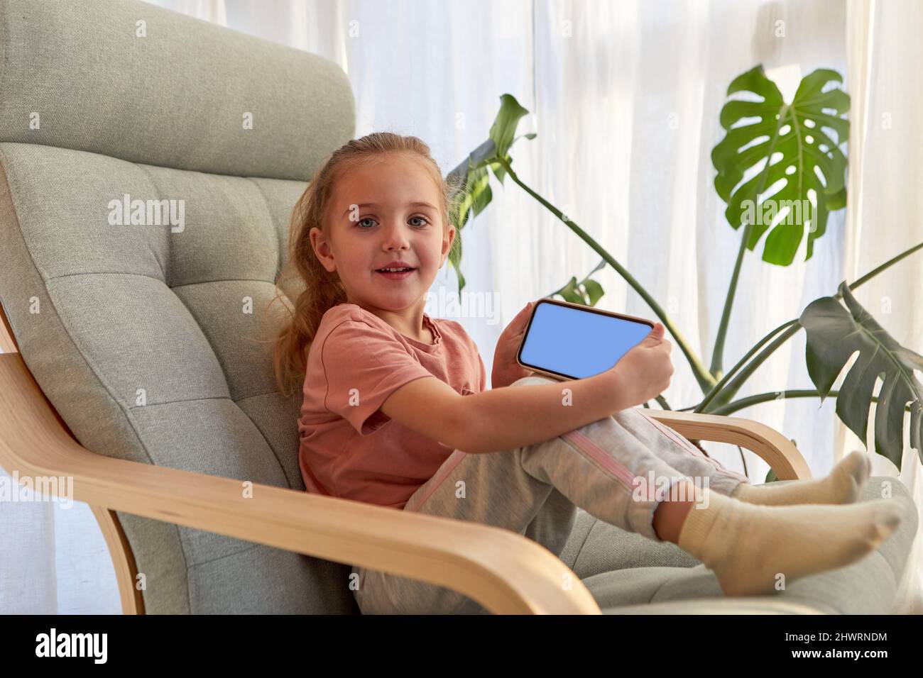 Vista laterale di adorabile bambina con lunghi capelli biondi in abiti casual sorridente e guardando la macchina fotografica mentre si siede su una comoda poltrona e demone Foto Stock