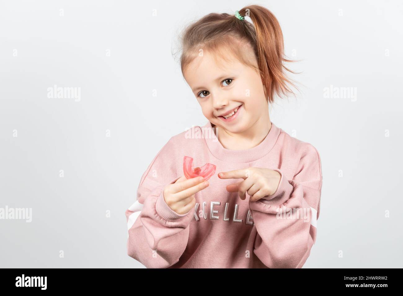 Carino ragazza caucasica piccola con i capelli biondi sta tenendo un addestratore dentario di miofunctional rosa sul bianco lo sfondo. Tariner dentale è fatto per aiutare equala Foto Stock