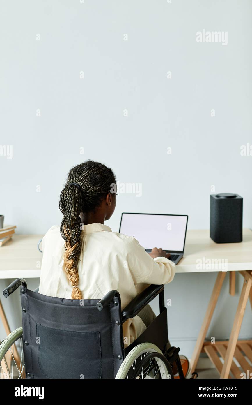 Vista posteriore minima della giovane donna afroamericana in sedia a rotelle utilizzando un computer portatile mentre si lavora in ufficio con dispositivi intelligenti accessibili Foto Stock