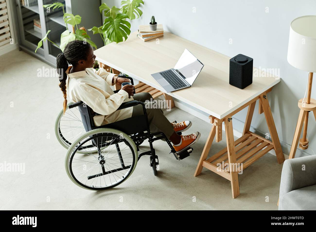 Vista ad alto angolo della giovane donna afroamericana in sedia a rotelle utilizzando un computer portatile mentre si lavora in ufficio con dispositivi intelligenti accessibili Foto Stock