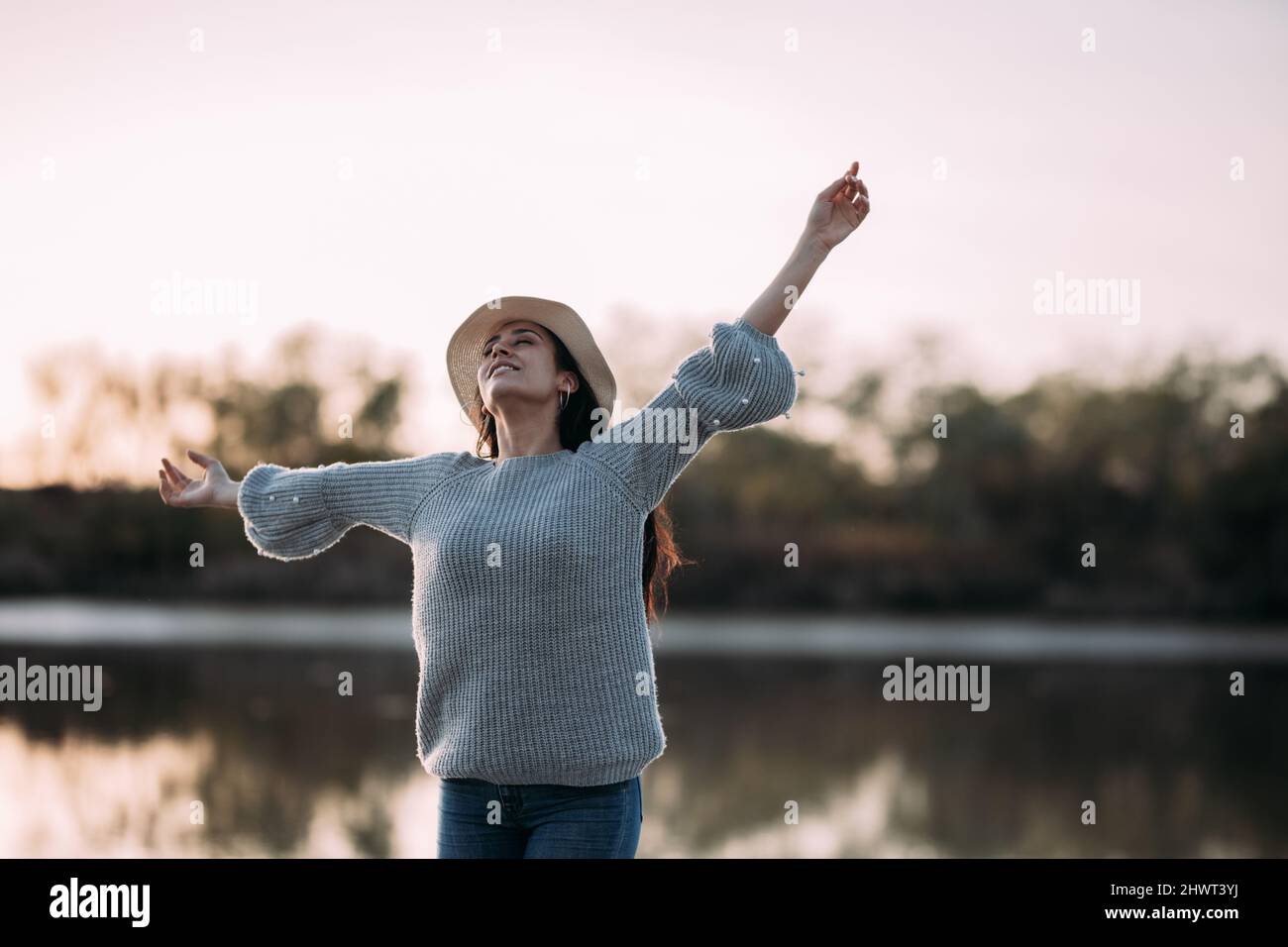 Donna brunetta che si sente libera sulla riva di un fiume con un cappello che alza le braccia Foto Stock