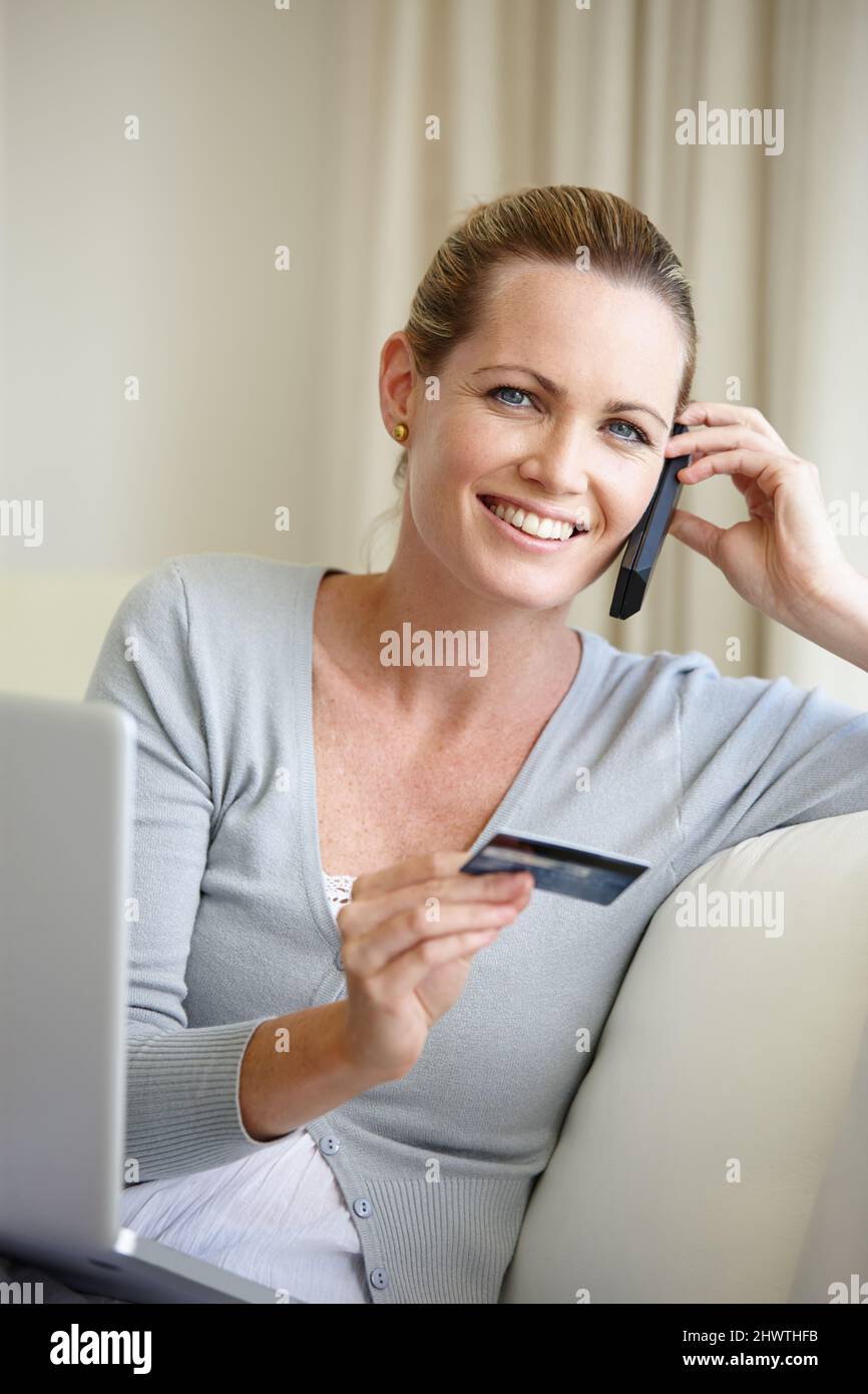 Tutto ciò che voglio a portata di mano. Una giovane donna che parla sul cellulare mentre tiene una carta di credito e si siede con un computer portatile. Foto Stock