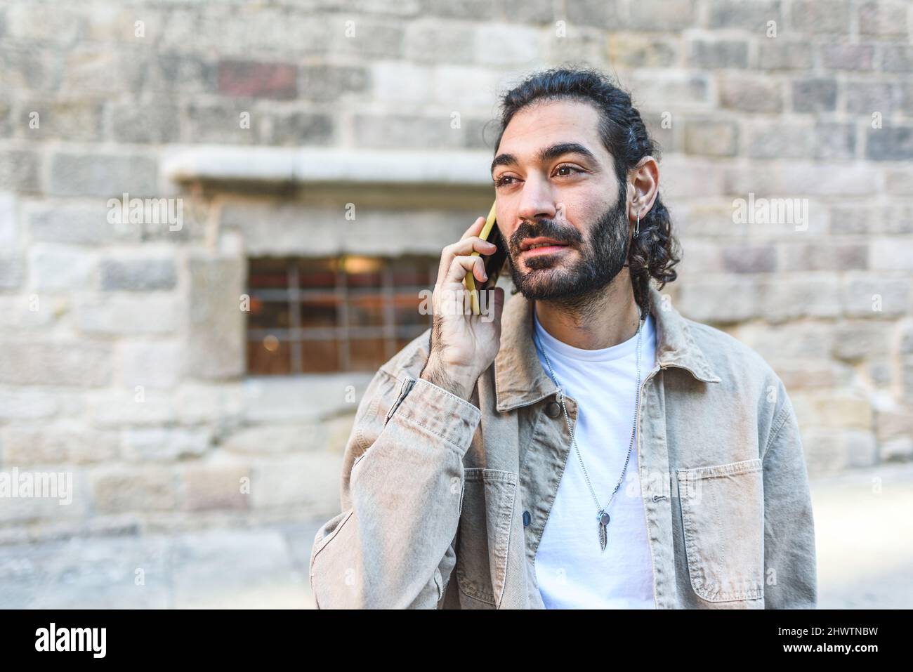 Turista maschio bearded con i capelli scuri che guardano via mentre hanno conversazione telefonica sulla strada vicino al vecchio edificio shabby in città Foto Stock