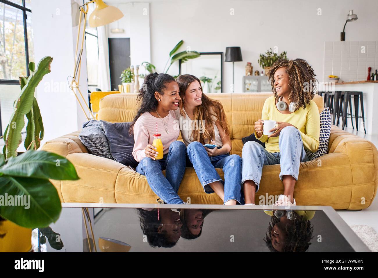 Tre allegre amiche sorridenti e divertenti a casa - donne divertenti che festeggiano la seduta sul divano del soggiorno bevendo succo di frutta e caffè Foto Stock