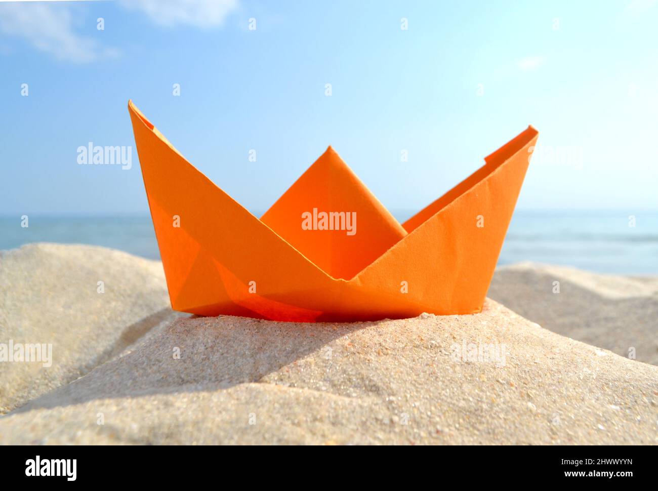 Barca di carta arancione brillante in sabbia gialla sullo sfondo delle onde del mare e del cielo blu Foto Stock