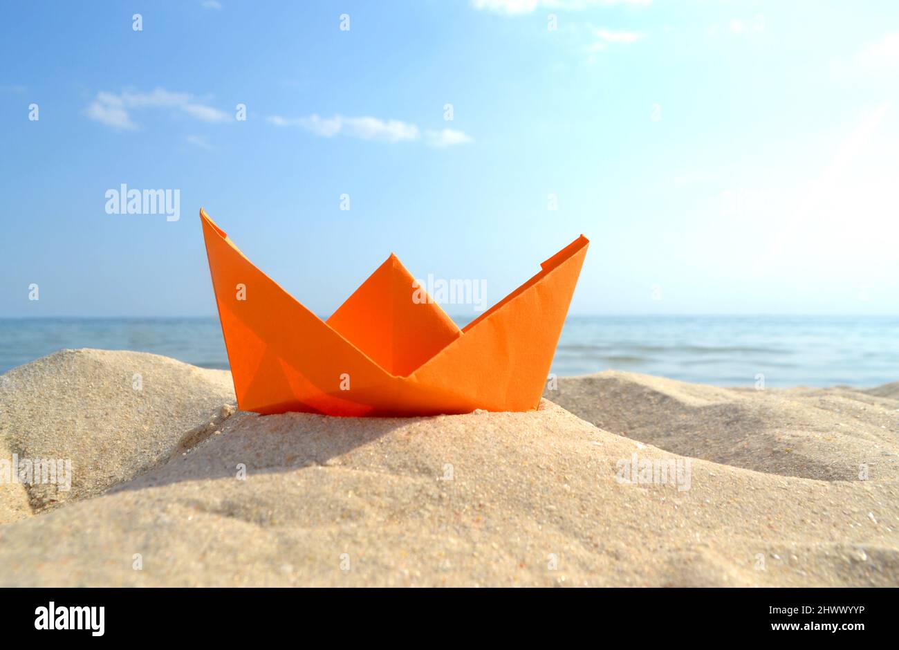 Barca di carta arancione brillante in sabbia gialla sullo sfondo delle onde del mare e del cielo blu Foto Stock