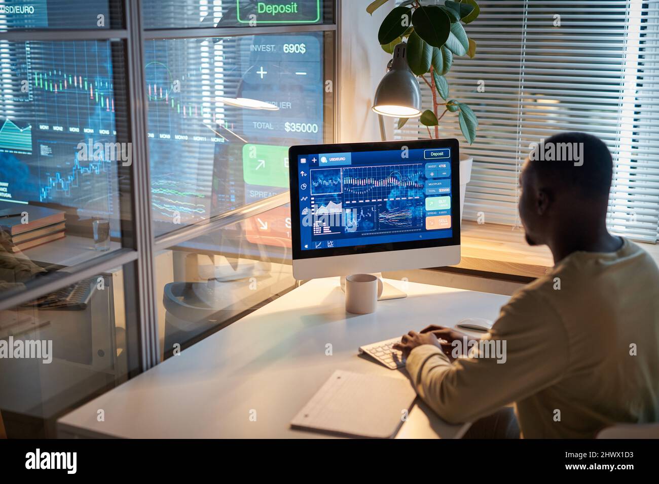 Vista posteriore del programmatore africano seduto sul posto di lavoro di fronte al computer e lavorando con i grafici sul monitor, lavora fino a tarda notte Foto Stock