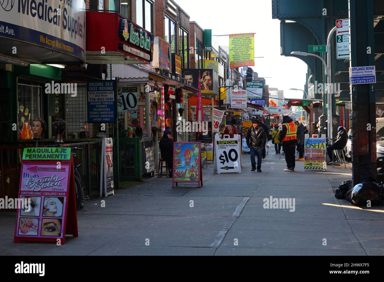 Una persona che indossa una camicia American Flag e un cappello di New York City cammina lungo Roosevelt Ave circondato da segni di lingua spagnola. Jackson Heights, Queens. Foto Stock