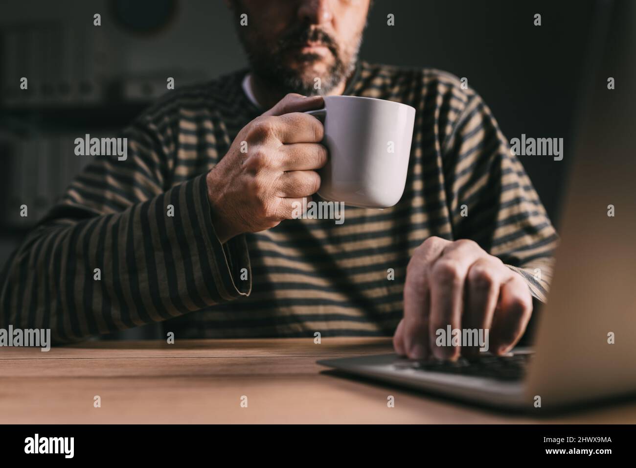 Freelance indipendente che lavora su un computer portatile in ufficio a casa a tarda notte e beve caffè per rimanere svegli, fuoco selettivo Foto Stock