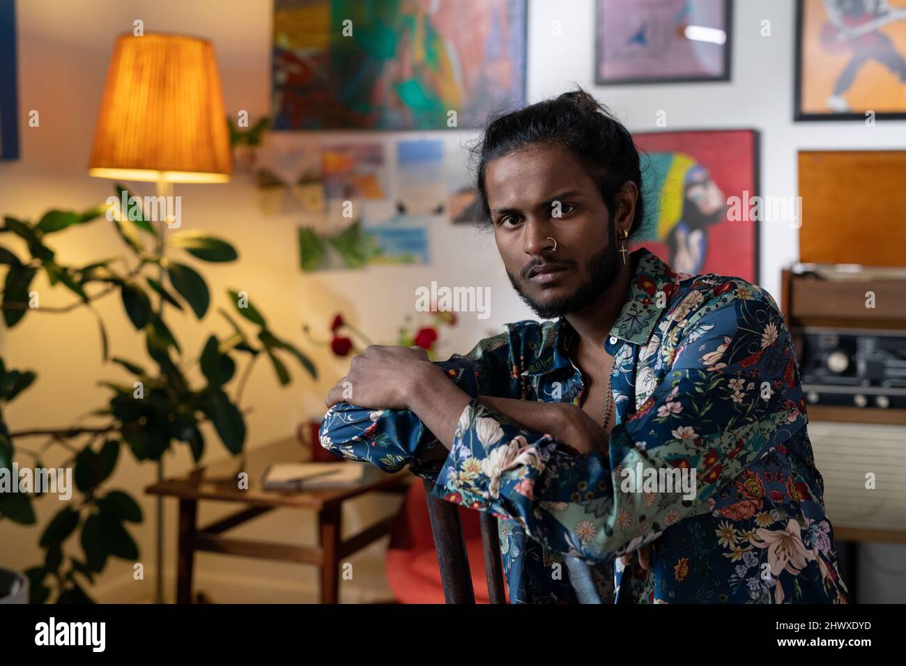 Giovane uomo sereno di etnia indiana che indossa una camicia elegante seduta in studio d'arte o di design di fronte alla macchina fotografica contro dipinti sulle pareti Foto Stock