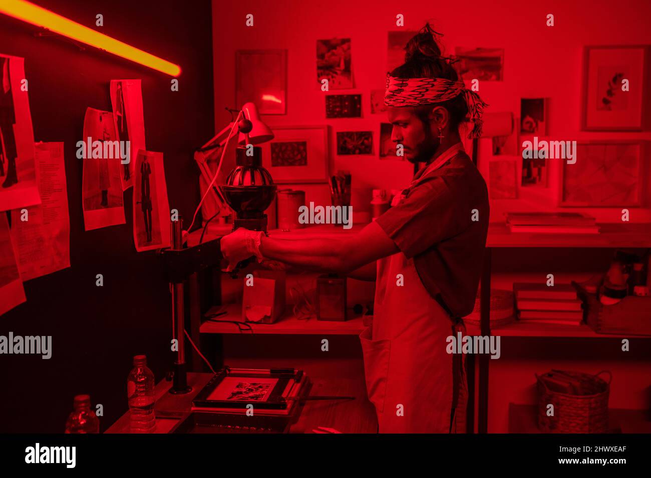 Giovane fotografo maschile serio che usa la foto ingrandisci mentre fa fotografie in bianco e nero in laboratorio rosso illuminato Foto Stock