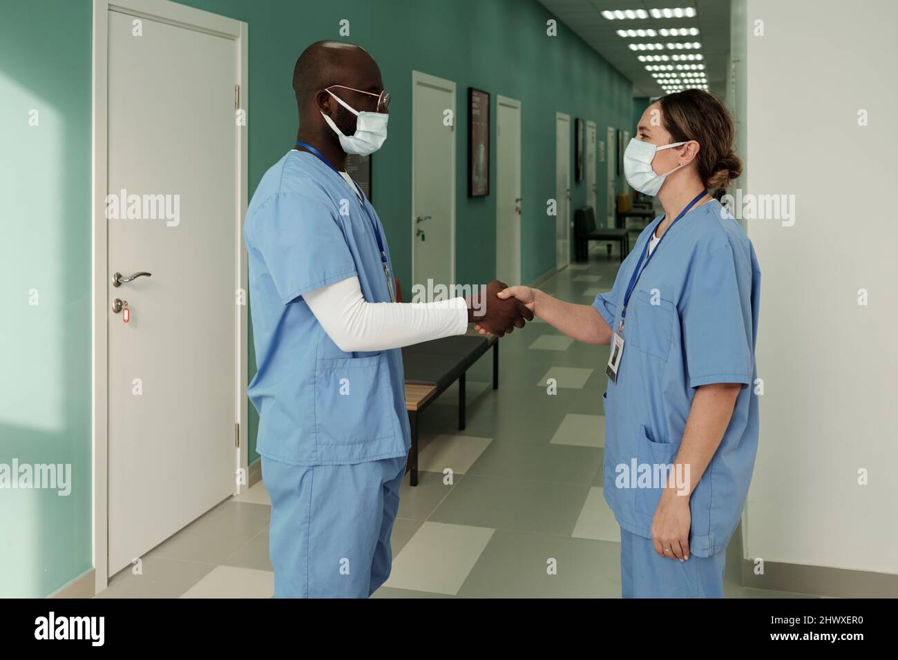 Due giovani professionisti generali interrazziali di successo in scrub medici handshaking nel corridoio dell'ospedale contemporaneo Foto Stock