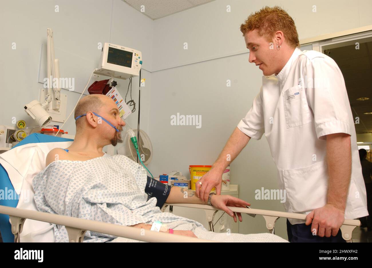 Un malato di attacco di cuore maschio si deposita nel suo letto con una scorta di ossigeno come un infermiere maschio parla con lui.(MODELLO RILASCIATO) Foto Stock