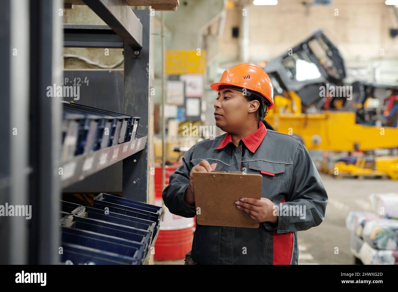Lavoratrice femminile di impianti industriali che fa note di revisione nel documento mentre guarda attraverso diversi contenitori di plastica con parti di ricambio Foto Stock