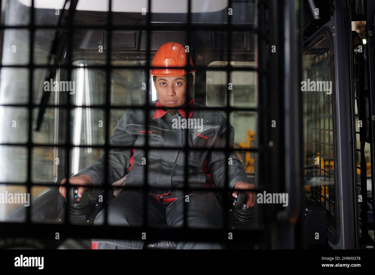 Lavoratrice femminile in elmetto e abbigliamento da lavoro che tiene i cambi marcia mentre guida la macchina da costruzione e guarda in avanti Foto Stock