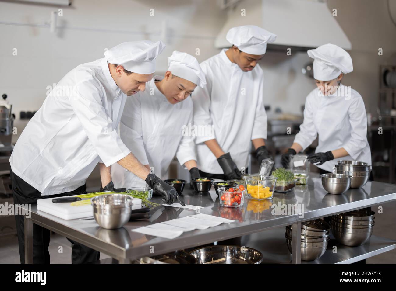 Gruppo di cuochi multirazziale che mescola gli ingredienti per portare via il cibo in cucina professionale. Concetto di cucina scura per cucinare per la consegna. Idea di lavoro di squadra in un ristorante. Persone diverse al lavoro Foto Stock