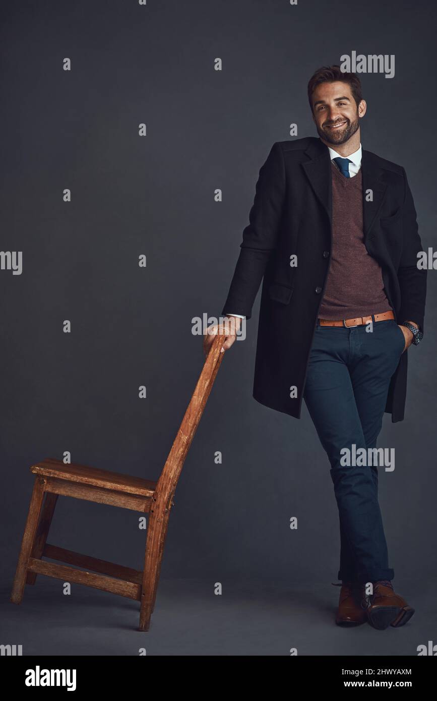 Indossa il tuo meglio e senti il tuo meglio. Ritratto di studio di un giovane uomo vestito con eleganza in piedi accanto ad una sedia su sfondo grigio. Foto Stock