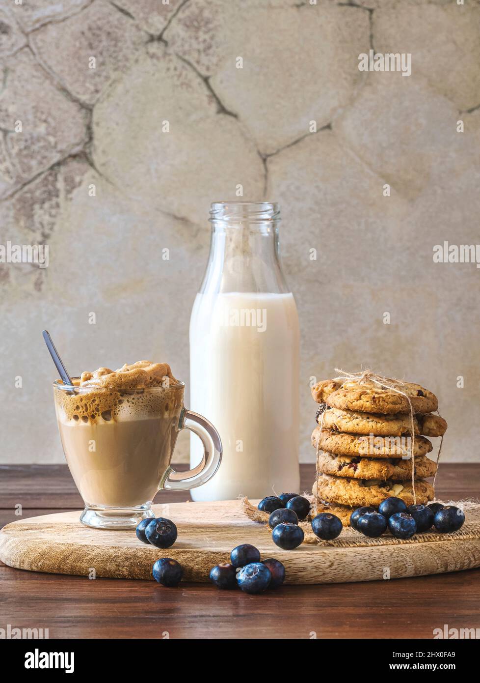 Caffè Dalgona in un bicchiere trasparente con biscotti al mirtillo e mirtilli su un asse di legno. Sfondo rustico parete. Caffè coreano molto alla moda. Foto Stock